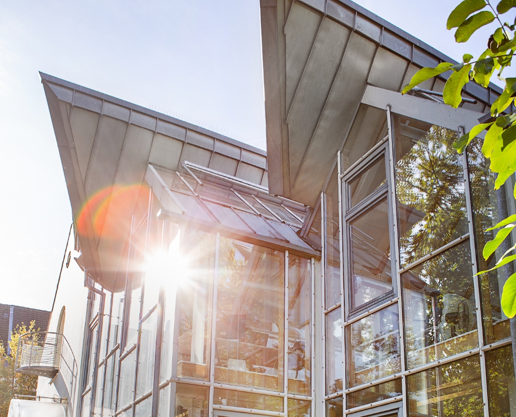 Modernes Gebäude mit Glasfront reflektiert Sonnenlicht, umgeben von Bäumen