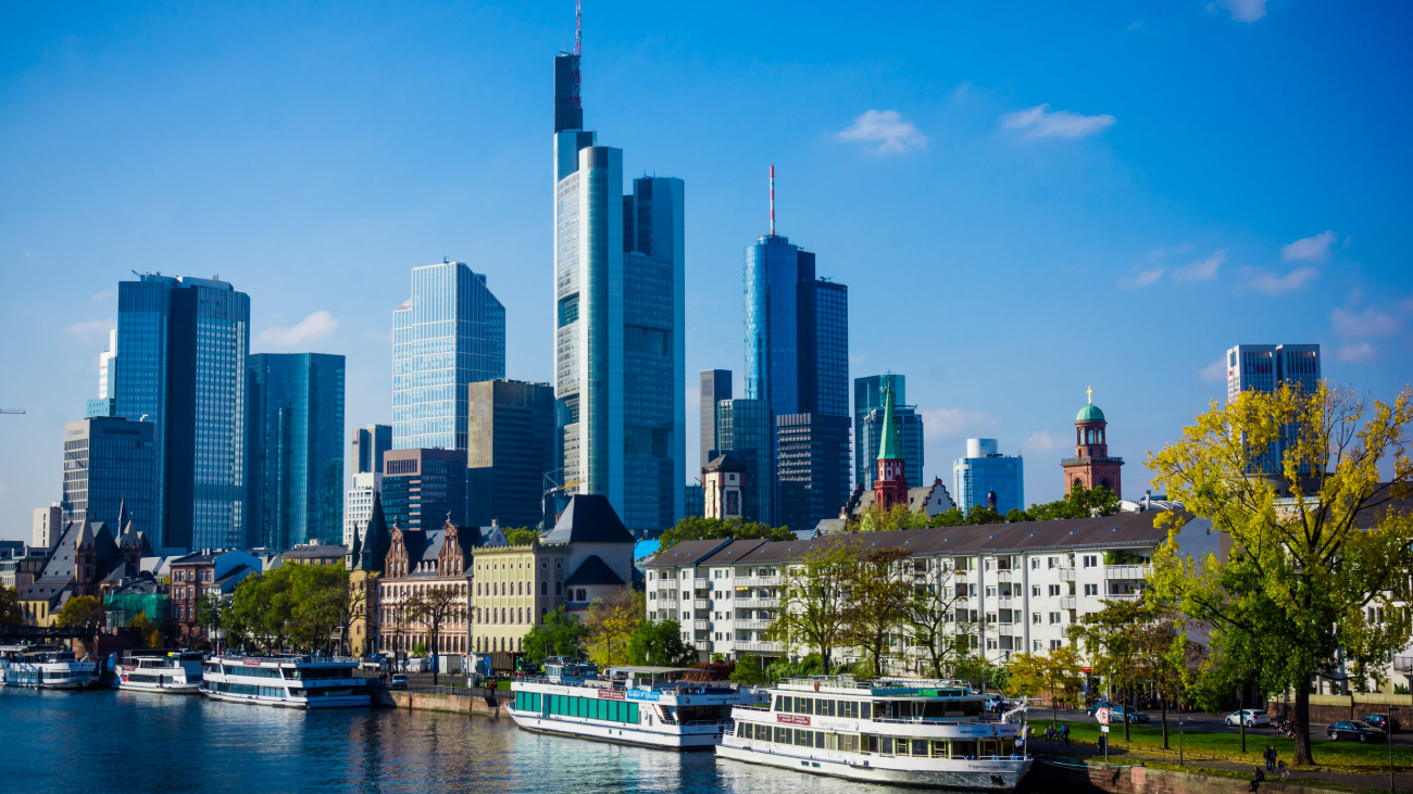 Lohnabrechnung Frankfurt: Hochhäuser reflektieren Sonnenlicht, Boote fahren auf Fluss im Stadtbild