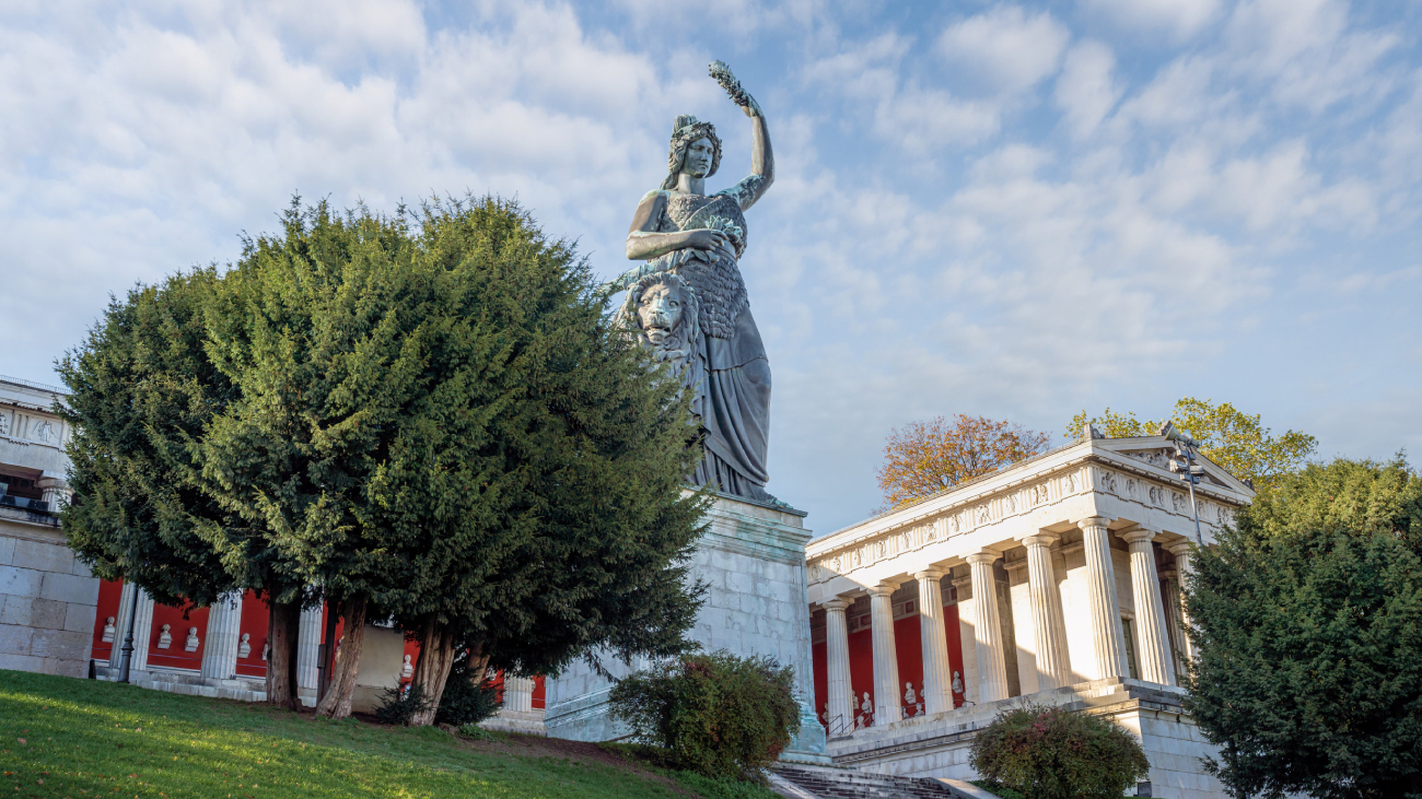 Lohnabrechnung München: Statue steht neben Bäumen, Gebäude mit Säulen im Hintergrund